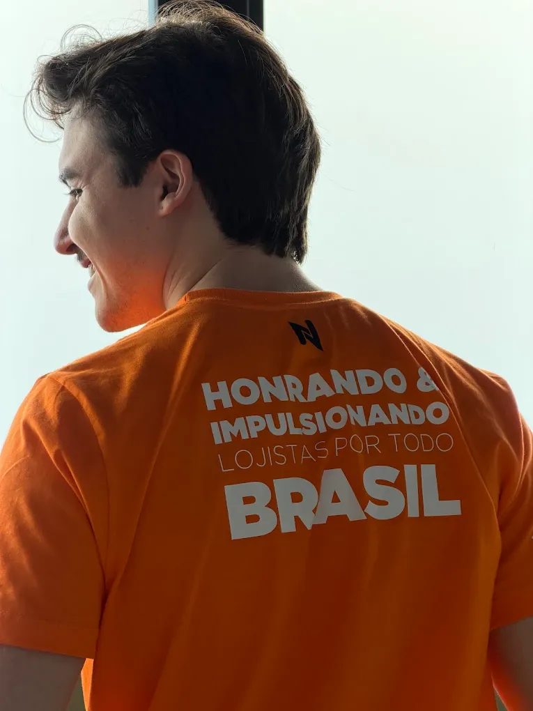 Integrante da equipe de costas, com a camiseta laranja da Novvacar escrita "Honrando e impulsionando lojistas por todo Brasil"
